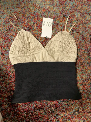 Beige och svart linnetopp från Zara - Trendig linnetopp från Zara med smala axelband. Överdelen är beige med rynkade detaljer och v-ringning, medan nederdelen är svart och ribbad. Perfekt för en stilren och modern look.