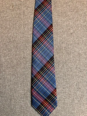 Rutmönstrad slips från Röda Sigillet - Snygg slips i blått, rött och svart med rutigt mönster från Röda Sigillet. Tillverkad i ett glansigt tyg som ger en stilren look. Perfekt accessoar för att lyfta din outfit med lite färg och mönster. Svensk tillverkad 