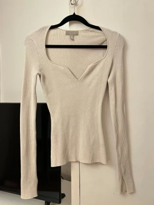Ribbad beige topp från H&M - Superfin ribbad topp från H&M i beige med lång ärm och snygg v-ringning. Toppen är figurnära och har en mjuk, stretchig känsla som passar perfekt till jeans eller kjol. Enkel men ändå trendig design som är lätt att matcha.
