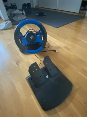 Ratt och pedaler till spelkonsol - Säljer en Thrustmaster ratt och pedaler för spelkonsol. Ratten är svart och blå med ergonomiskt grepp och flera knappar. Pedalerna är svarta och kopplas enkelt ihop med ratten via kabel. Perfekt för racing- och bilspel.