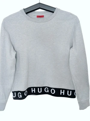 Grå sweatshirt från Hugo Boss Unik Slutsåls - Snygg grå sweatshirt från Hugo Boss med svart resår nertill där det står 'HUGO' i vitt. Tröjan har rund halsringning och långa ärmar. Perfekt för en avslappnad och trendig stil. Nyskick 