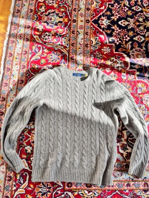 Grå kabelstickad tröja Polo Ralph Lauren - Kabelstickad grå tröja från Polo Ralph lauren tröja i lammull. Klassisk design med diskret broderad logga på bröstet. Perfekt för lager-på-lager och enkel att matcha med jeans eller kjol. OBS litet hål i högerarm men enkelt att fixa 