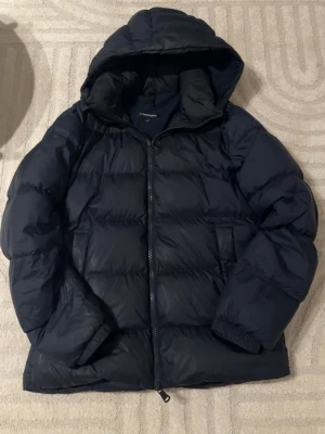 Mörkblå dunjacka från J.Lindeberg - Snygg mörkblå dunjacka från J.Lindeberg med huva finns enstaka skada på ena fickan se bild överlag väldigt fin!