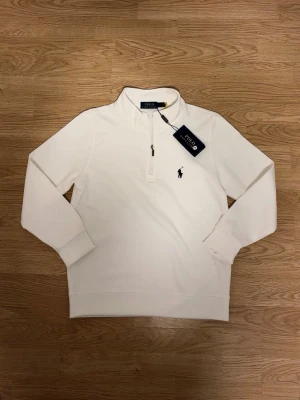 Vit quarter zip från Polo Ralph Lauren - Snygg vit långärmad tröja från Polo Ralph Lauren med quarter zip och klassisk broderad logga på bröstet. Tröjan har hög krage och är tillverkad i mjukt material som känns skönt mot huden. Perfekt för en clean och stilren look.