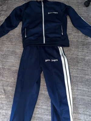 Storlek s passar i M också. Mörkblå tracksuit från Palm Angels - Storlek s passar i m också. Snygg mörkblå tracksuit från Palm Angels med  vita detaljer. Setet består av en zip-jacka med vita ränder längs ärmarna och matchande byxor med vita sidoränder. Båda delarna har Palm Angels-loggan tryckt i vitt. Perfekt för sport eller chill.
