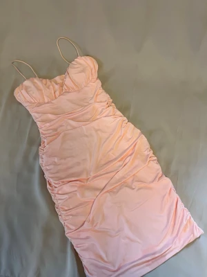 Ljusrosa klänning med smala band - Supersöt ljusrosa kortklänning med smala axelband. Klänningen har rynkade sidor som ger en snygg form och är tillverkad i ett mjukt, stretchigt material. Knälång. 