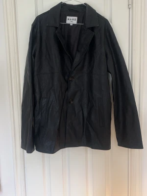 Svart skinnjacka från NA-KD - Stilren svart skinnjacka från NA-KD med klassisk krage och tre knappar framtill. Jackan har raka ärmar och två diskreta fickor på sidorna. Perfekt för dig som gillar en tidlös och clean look.