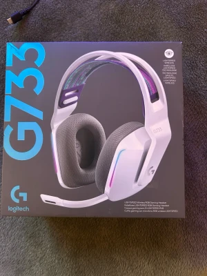 Logitech G733 trådlöst gamingheadset - Snyggt vitt Logitech G733 gamingheadset med RGB-belysning och mjuka grå öronkuddar. Trådlös modell med avtagbar mikrofon och justerbart huvudband i färgglatt tyg. Perfekt för gaming med USB-laddning och LIGHTSPEED-teknologi.
