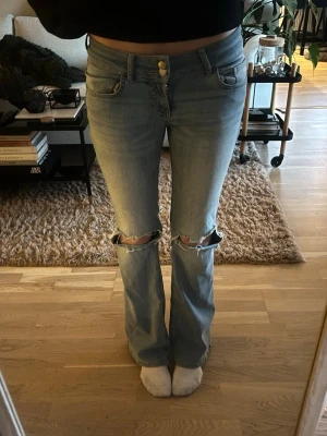 Ljusblå slitna bootcut jeans - Säljer ett par ljusblå bootcut jeans med slitningar över knäna och knapp fickor på baksidan. Jeansen är i bra skick och kommer från Nelly! De är i storlek 36 och innerbenslängden är 82cm och midjemåttet är 74cm.
