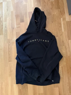 Svart hoodie från Tommy Jeans - Snygg svart hoodie från Tommy Jeans med logga broderad över bröstet. Klassisk modell med huva och dragsko, samt ribbade muddar vid ärmslut och nederkant. Perfekt för en avslappnad och trendig stil. Använd fåtal gånger.