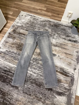 Grå raka jeans från Levi's - Säljer ett par snygga grå jeans från Levi's med raka ben och klassisk femficksdesign. Jeansen har en ljus tvätt och subtila slitningar framtill för en avslappnad look. Perfekta till vardags och lätta att matcha med olika stilar. Jeansen har en liten defekt som jag har fixat i vänster ficka och en liten defekt i bak lapp som inte syns så mycket 