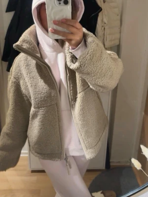 Beige teddyjacka med dragkedja - Mysig beige teddyjacka med hög krage och dragkedja framtill. Jackan har en boxig passform och är tillverkad i fluffigt teddy-material som ger en trendig och avslappnad look. Perfekt för lager-på-lager-stil under kyliga dagar.