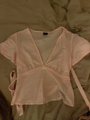 Ljusrosa omlottopp med knytband - Supersöt ljusrosa omlottopp med korta ärmar och knytband i midjan. Toppen har v-ringning och är tillverkad i ett mjukt bomullsmaterial. Perfekt för en trendig och avslappnad look.