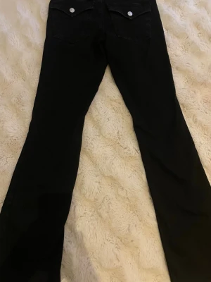 Svarta bootcut jeans med fickor - Säljer ett par svarta bootcut jeans, silverfärgade knappar bak. Jeansen har en rak passform upptill och lätt utsvängda ben. Perfekta till en avslappnad och trendig look.