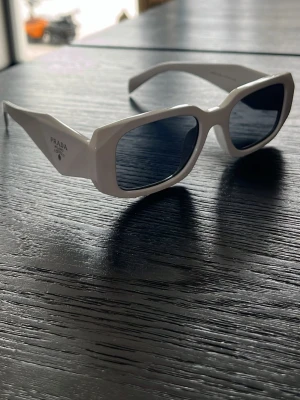 Vita rektangulära solglasögon Prada - Snygga vita solglasögon från Prada med rektangulär form och breda skalmar. Glasen är mörka och bågarna har en modern, chunky design med Prada-loggan på sidan. Perfekt accessoar för att lyfta din stil.