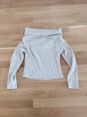 Ljusgrå offshoulder topp - Trendig ljusgrå offshoulder topp med långa ärmar och figurnära passform. Tillverkad i mjukt bomullsmaterial som känns skönt mot huden. Perfekt för dig som vill ha en stilren och enkel look med en twist.
