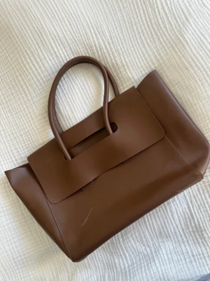 Brun handväska - Snygg brun handväska i slätt konstläder med stilrena linjer och två handtag. Väskan har en enkel, modern form och ett stort lock framtill. 