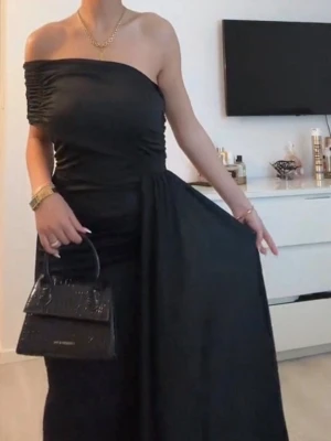Svart offshoulder långklänning - Elegant svart långklänning med offshoulder-design och figurnära passform. Klänningen har en draperad överdel och en lång, svepande kjol som ger en lyxig känsla. Perfekt för dig som vill sticka ut på nästa stora event.