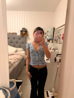 Blå blommig croppad blus med volang - Säljer en blå och vit blommig croppad blus med volangkant och v-ringning. Toppen har korta volangärmar och smockad midja som ger en snygg passform. Perfekt för dig som gillar trendiga och feminina plagg.