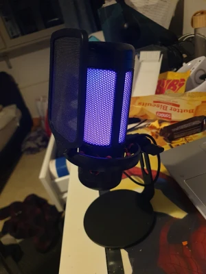 USB-mikrofon med RGB-belysning - Säljer en svart USB-mikrofon med coolt blått RGB-ljus och popfilter. Perfekt för streaming, gaming eller podd. Mikrofonen står på ett stabilt stativ och har en modern, rund design i metall och plast.