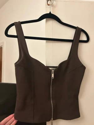 Brun croppad topp med dragkedja - Snygg brun croppad topp med breda axelband och dragkedja framtill. Toppens form är figurnära och den är tillverkad i ett stretchigt material som känns bekvämt. Perfekt att styla med jeans eller kjol för en trendig look.