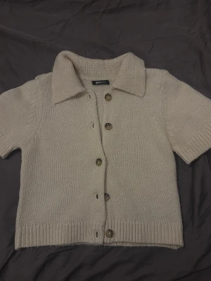 Beige stickad kortärmad kofta - Stickad beige kofta från Gina Tricot med korta ärmar, knappar framtill och klassisk krage. Mjuk och mysig, perfekt för lager-på-lager. Ribbstickad nederkant och ärmslut ger en snygg finish.