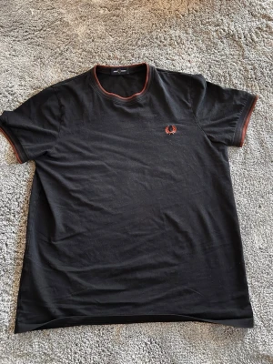 Svart Fred Perry t-shirt XS - Svart t-shirt från Fred Perry med brunt broderat logomärke på bröstet och bruna detaljer vid hals och ärmslut. Klassisk passform och korta ärmar. Perfekt för en stilren och enkel look.
