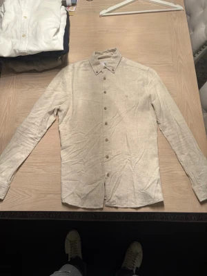 Beige linneskjorta med knappar - Stilren beige skjorta i linne med klassisk krage och knäppning framtill. Skjortan har lång ärm och en avslappnad passform, perfekt för en fräsch och enkel look. Passar dig som gillar naturliga material och tidlös stil.