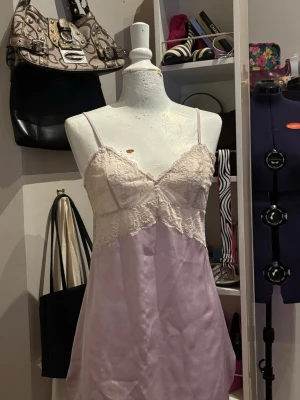 Ljuslila spetsklänning / nattlinne - Romantisk ljuslila klänning/nattlinne från Ella M i satinliknande tyg med vackra spetsdetaljer över bysten. Den feminina designen tillsammans med de tunna axelbanden ger ett elegant och mjukt uttryck. Baksidan har en dekorativ knytning som ger en fin detalj och justerbar känsla. Perfekt som nattlinne, loungewear eller en romantisk sommarklänning.