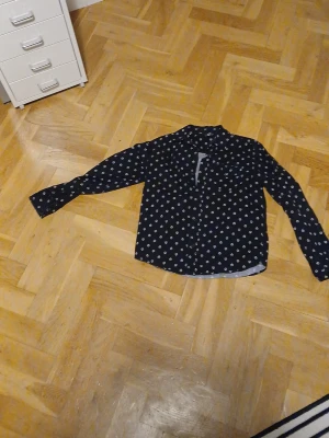 Svart skjorta med döskallemönster - Cool svart skjorta med vita döskallar över hela tyget. Skjortan har lång ärm, klassisk krage och knäppning framtill. Perfekt för dig som vill sticka ut med en edgy stil.