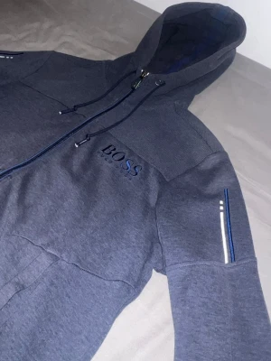 Hugo boss  - Snygg grå hoodie från BOSS med dragkedja och huva. Logotypen är broderad på bröstet och ärmarna har diskreta blå och vita detaljer. Perfekt för en avslappnad och cool stil. Materialet är mjukt och bekvämt. armbågarna har tappat lite tyg men de märks inte 