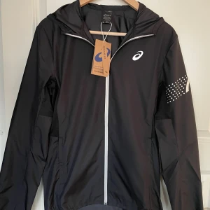 Svart löparjacka från Asics - Svart löparjacka från Asics med huva och hel dragkedja framtill. Jackan har vita detaljer, bland annat logga på bröstet och prickigt mönster på ena ärmen. Lätt och smidig modell i syntetmaterial, perfekt för löpning och träning utomhus.