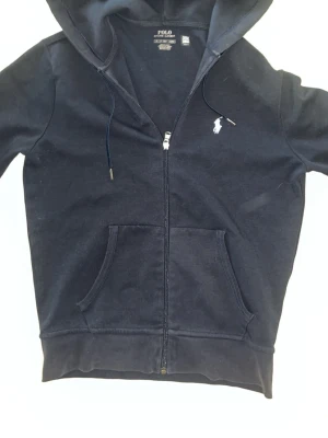 Mörk blå hoodie från Polo Ralph Lauren -  SER SLUTSIG UT MEN DET ÄR LJUSET!!! Mörk blå hoodie med dragkedja från Polo Ralph Lauren. Klassisk design med vit broderad logga på bröstet, huva med dragsko och känguruficka framtill. Tillverkad i mjuk bomull för en skön och avslappnad stil.