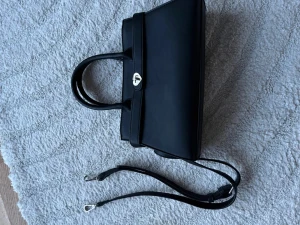 Svart handväska med hjärtspänne - Stilren svart handväska i slätt material med dubbla handtag och avtagbar axelrem. Väskan har ett unikt silverfärgat hjärtformat spänne framtill som detalj. Perfekt för dig som gillar minimalistisk design med en söt twist.