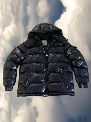 Svart glansig moncler maya dunjacka - Säljer en svart, glansig dunjacka från Moncler med huva och dragkedja framtill. Jackan har två snedställda fickor med dragkedja och klassisk pufferdesign. Perfekt för kalla vinterdagar och riktigt snygg streetstil.