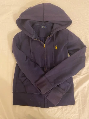 Mörkblå hoodie från Polo Ralph Lauren - Mörkblå hoodie från Polo Ralph Lauren med dragkedja framtill och gul broderad logga på bröstet. Klassisk huva och fickor på sidorna. Tillverkad i mjuk bomullsblandning som är skön att ha på sig. Perfekt för en avslappnad och stilren look.