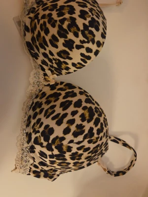 Leopardmönstrad push-up bh med spets - Snygg push-up bh med leopardmönster i beige, svart och guldiga toner. Bh:n har vadderade kupor och dekorativ spets längs kanterna samt justerbara axelband. Perfekt för dig som vill ha en djärv och trendig look. Priset kan diskuteras!