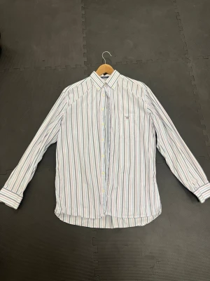 GANT Skjorta  - Snygg skjorta från Gant med tunna vertikala ränder i blått, rosa och grönt på vit botten. Klassisk krage, bröstficka med broderad logga och långa ärmar med manschettknappar. Perfekt för en stilren look.