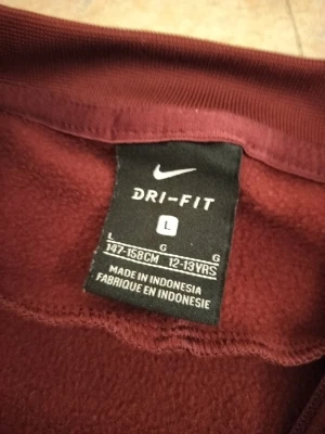 Vinröd Nike Dri-Fit träningströja - Säljer en vinröd långärmad träningströja från Nike med Dri-Fit material. Tröjan har coola orange detaljer längs ärmarna och är tillverkad i ett funktionsmaterial som transporterar bort fukt. Perfekt för träning eller sportaktiviteter. Barcelona 2018 kit.