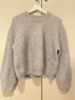 Grå ribbstickad tröja - Mysig grå stickad tröja med bred ribbstickning och rund hals. Tröjan har en avslappnad passform, långa ärmar och ribbade muddar vid ärmslut och nederkant. Perfekt för kyliga dagar och enkel att matcha med olika outfits.