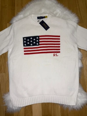 Vit stickad tröja Polo Ralph Lauren - Vit stickad tröja från Polo Ralph Lauren med amerikansk flagga i rött, vitt och blått på bröstet. Tröjan är ganska stor i storleken så jag hade nog sagt att storlek L är bäst för denna. Så det är en M men sitter som L.