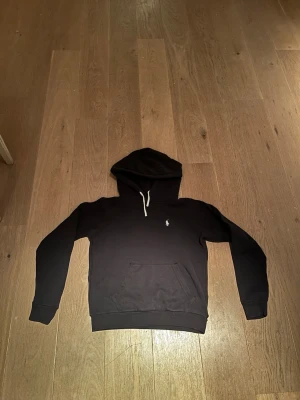 Svart hoodie från Polo Ralph Lauren - Snygg svart hoodie från Polo Ralph Lauren med klassisk vit logga broderad på bröstet. Hoodien har en stor huva med vita dragsnören och en praktisk magficka framtill. Tillverkad i mjukt bomullsmaterial som är skönt att bära.