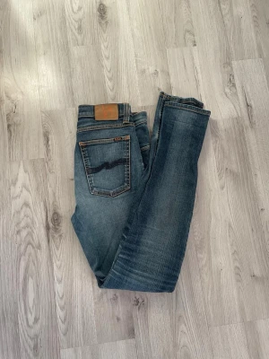 Blå jeans från Nudie Jeans - Priset är inte rispat i sten. Snygga blå jeans från Nudie Jeans med klassisk femficksdesign och kontrastsömmar. Jeansen har en tvättad look med slitningar och Nudie-loggan på bakfickan samt läderpatch i midjan. Perfekta för en avslappnad och trendig stil.