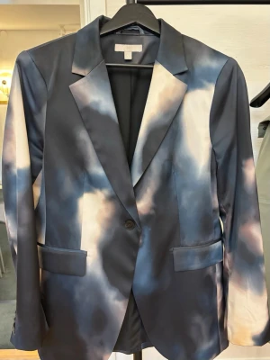 Blå tie-dye kavaj från H&M - Snygg kavaj från H&M i storlek 40 med unikt blått och vitt tie-dye mönster. Klassisk modell med slag, enkel knappstängning och två fickor framtill. Tillverkad i ett slätt, vävt material som ger en modern och cool look.