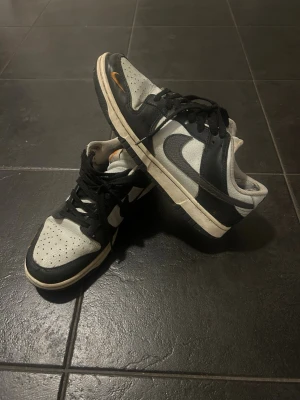 Nike Dunk Low svart/vit sneaker - Säljer ett par Nike Dunk Low sneakers i svart och vitt med klassisk Swoosh på sidan. Skorna har svart läderöverdrag, vit mesh på tåboxen och svarta skosnören. Yttersulan är vit med svarta detaljer. Perfekta för dig som gillar streetwear och retrostil.