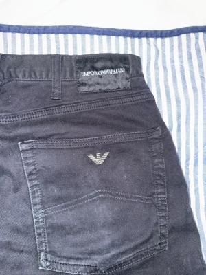 Svarta jeans från Emporio Armani - Snygga svarta jeans från Emporio Armani med klassisk femficksmodell och diskret logga på bakfickan. Jeansen har raka ben och är tillverkade i ett mjukt bomullsmaterial med lite stretch för extra komfort. Perfekta för en stilren och modern look. Knappen är lite lös men går nog att fixa hos en skräddare eller om man själv vet hur man gör.