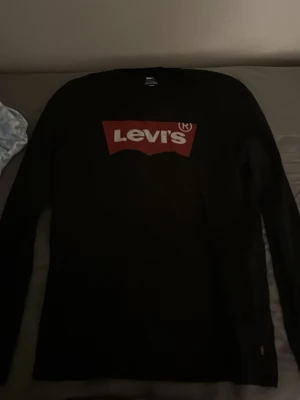 Svart långärmad topp från Levi's - Säljer en svart långärmad topp från Levi's med det klassiska röda och vita logotrycket framtill. Rund halsringning och mjukt bomullsmaterial som känns skönt mot huden. Perfekt för dig som gillar enkel och tidlös streetstil.