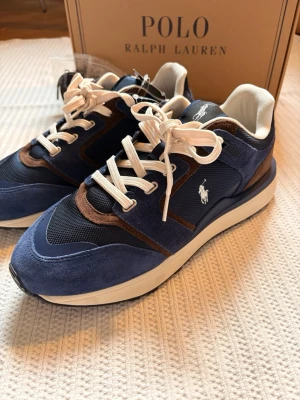 Ralph Lauren sneakers blå - Helt nya! Snygga sneakers i blå mocka och mesh från Polo Ralph Lauren med vita och bruna detaljer. Klassisk låg modell med vita snören och logga på sidan. Tjock sula för extra komfort. Perfekta för dig som vill ha stilrena och trendiga skor. Nypris 1799kr