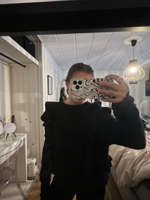 Svart volangtröja med puffärm - Cool svart tröja med volangdetaljer längs ärmarna och axlarna. Tröjan har puffiga ärmar som ger en trendig och edgy look. Perfekt för dig som vill sticka ut och addera lite attityd till outfiten.