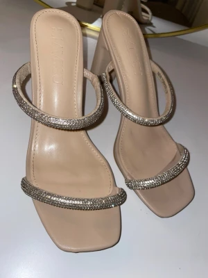 Beige pumps med strassdetaljer - Stilrena pumps i beige med fyrkantig tå och två remmar täckta av glittrande strass. Klackarna är höga och ger en elegant look. Perfekta för att lyfta din outfit med lite extra bling.